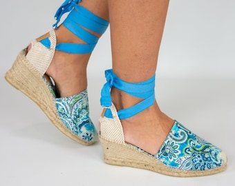 May include: Blue and green patterned wedge espadrille sandals with blue ribbon laces. The sandals have a woven jute platform and a wedge heel.