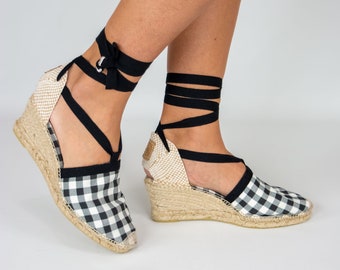 May include: Espadrille wedge sandals with a black and white gingham pattern. The sandals have a woven jute sole and black wrap-around ankle ties. The wedge heel provides lift and support. The overall design is a blend of classic and contemporary styles.