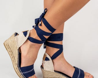 May include: Navy blue espadrille wedge sandals with woven jute soles and ankle ties. The open-toe sandals feature a navy blue fabric upper with white stitching and a beige canvas heel counter. The ankle ties wrap around the ankle and tie in a bow.