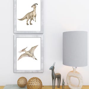May include: Two framed dinosaur art prints. One print features a Parasaurolophus, the other a Pterodactyl. The prints are in white frames. Decorative gold spheres, a gray lamp, and metal animal figurines sit on a wooden surface.