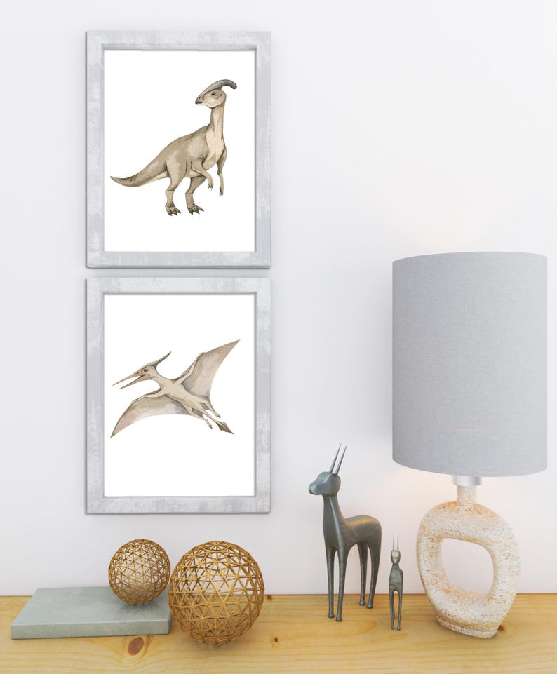 May include: Two framed dinosaur art prints. One print features a Parasaurolophus, the other a Pterodactyl. The prints are in white frames. Decorative gold spheres, a gray lamp, and metal animal figurines sit on a wooden surface.