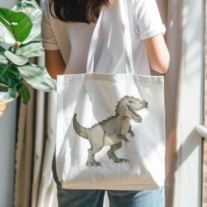 May include: A white canvas tote bag featuring a printed illustration of a grey Tyrannosaurus Rex dinosaur. The dinosaur is depicted in profile with its mouth open, showing sharp teeth, and has a textured, scaly appearance on its body.