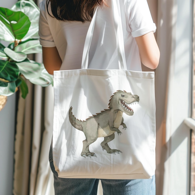 May include: A white canvas tote bag featuring a printed illustration of a grey Tyrannosaurus Rex dinosaur. The dinosaur is depicted in profile with its mouth open, showing sharp teeth, and has a textured, scaly appearance on its body.