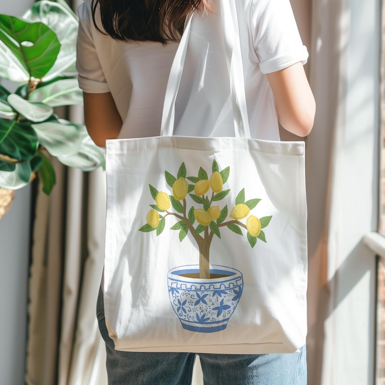 May include: White tote bag featuring a lemon tree illustration. The tree has yellow lemons, green leaves, and a brown trunk, planted in a blue and white patterned pot. The bag is carried by a person.