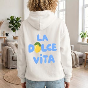 May include: White hoodie with the phrase "LA DOLCE VITA" in light blue letters, a yellow lemon graphic, and green leaves. The hoodie is made of a soft material and has a hood.