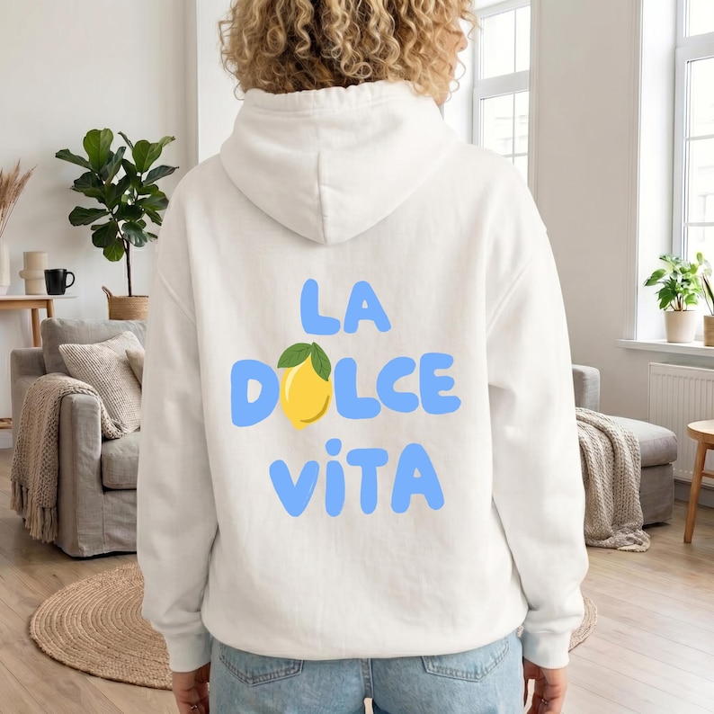 May include: White hoodie with the phrase "LA DOLCE VITA" in light blue letters, a yellow lemon graphic, and green leaves. The hoodie is made of a soft material and has a hood.