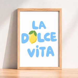 May include: A framed art print with the words "LA DOLCE VITA" in light blue, with a yellow lemon and green leaf replacing the "O" in "DOLCE". The frame is made of light brown wood. The print is on a white background.