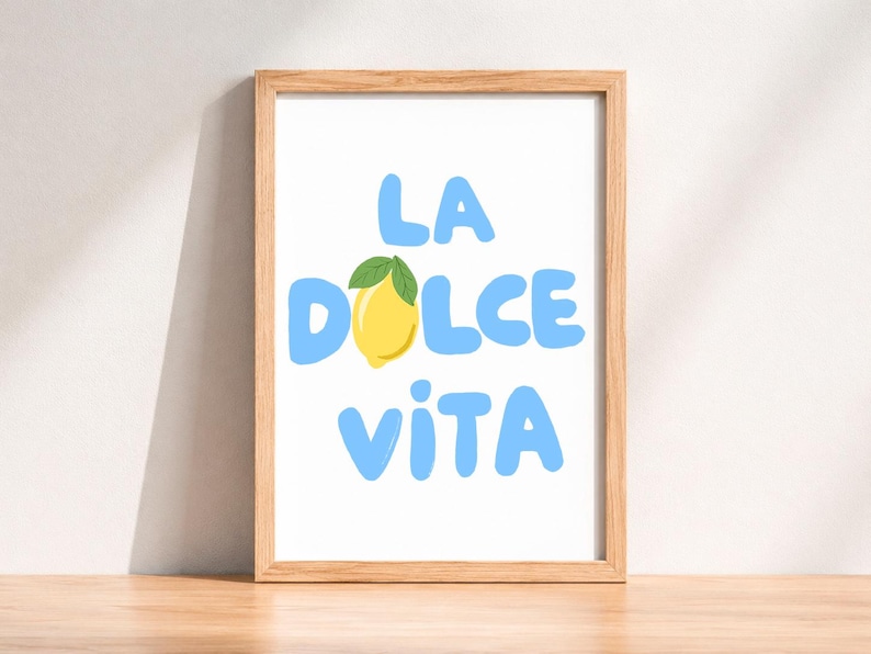 May include: A framed art print with the words "LA DOLCE VITA" in light blue, with a yellow lemon and green leaf replacing the "O" in "DOLCE". The frame is made of light brown wood. The print is on a white background.