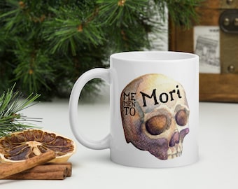 May include: White ceramic coffee mug with a watercolor skull illustration and the text "Memento Mori" in black lettering.