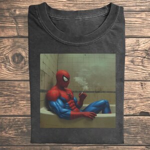 May include: A charcoal gray t-shirt featuring a graphic print of a red and blue costumed character sitting in a bathtub, holding a smoking cigarette. The character has a muscular build and is positioned against a tiled wall background.