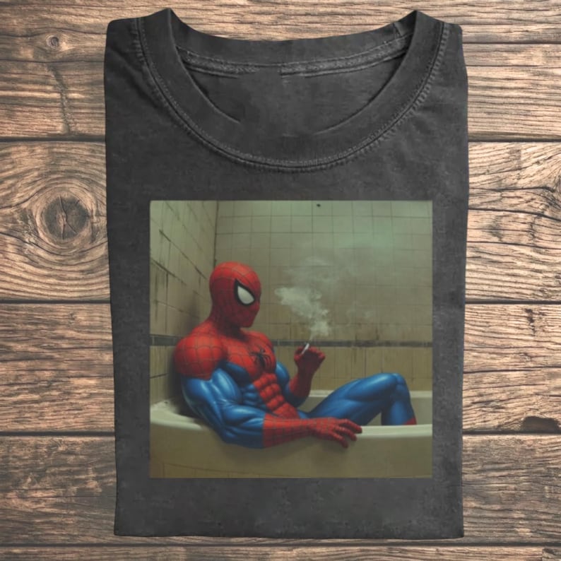 May include: A charcoal gray t-shirt featuring a graphic print of a red and blue costumed character sitting in a bathtub, holding a smoking cigarette. The character has a muscular build and is positioned against a tiled wall background.