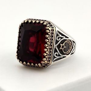 May include: A silver ring with a large, rectangular, deep red gemstone. The ring has a gold-colored bezel around the stone and decorative detailing on the sides, including a crescent moon and star design. The ring is set against a white background.