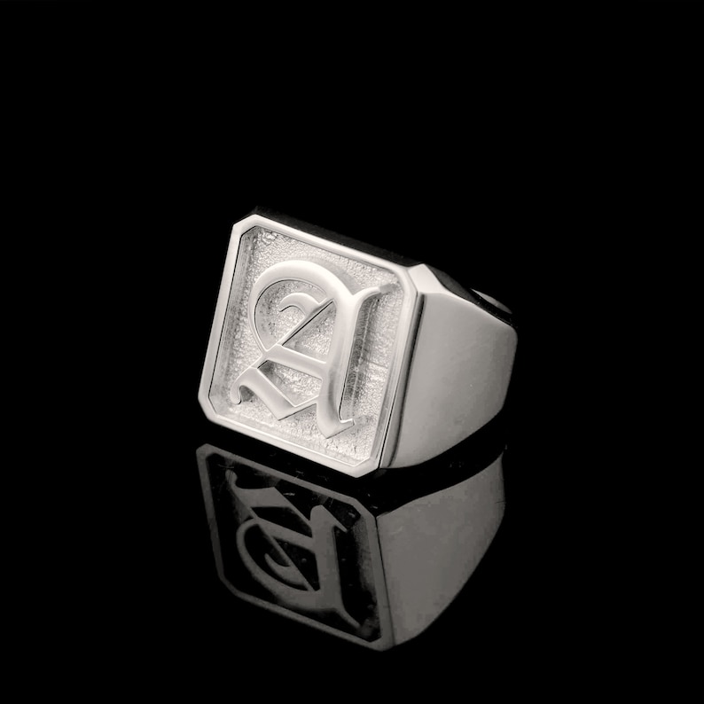 May include: A silver signet ring with a square face featuring a raised, stylized letter 'A' in a gothic font.