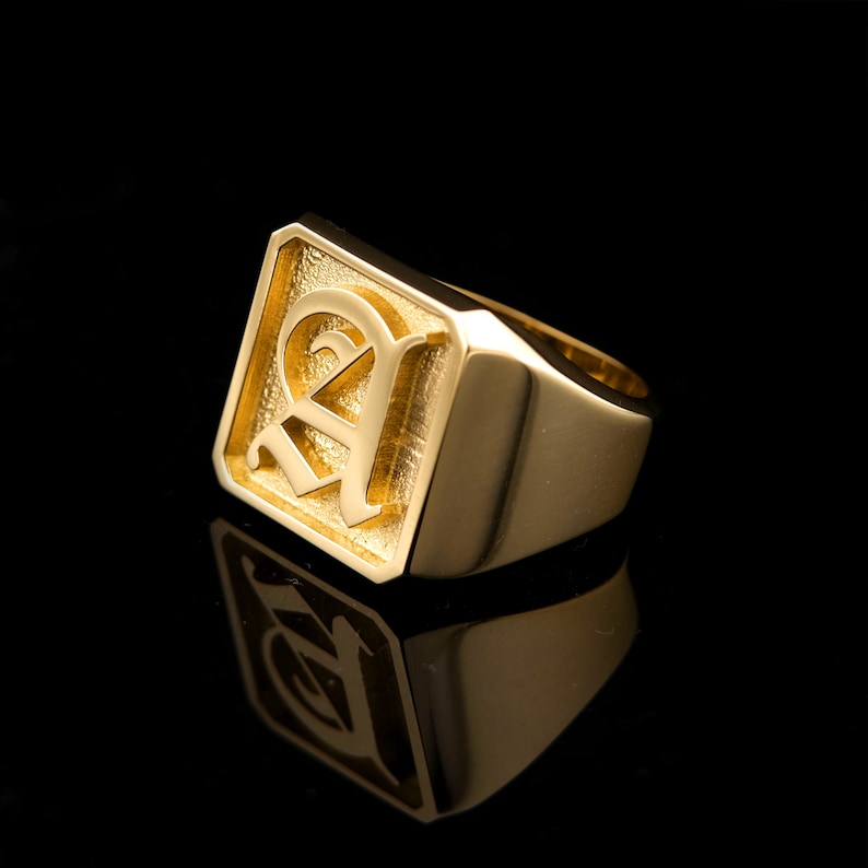 May include: A gold signet ring with a square face featuring a raised, stylized letter 'A' in a script font.