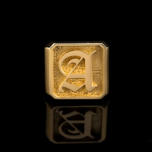May include: A gold signet ring with a raised, textured, old English letter 'A' in the center.