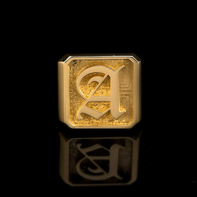 May include: A gold signet ring with a raised, textured, old English letter 'A' in the center.