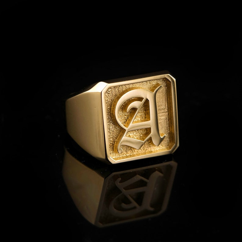 May include: A gold signet ring with a square face featuring a raised, stylized letter 'A' in a gothic font.