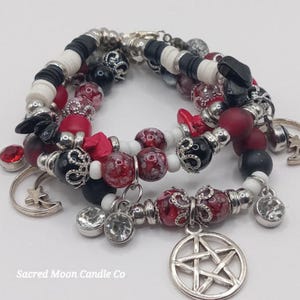 May include: A stack of beaded bracelets with charms. The bracelets feature red, black, white, and silver beads. Charms include a pentagram, crescent moon, and crystal pendants. The text "Sacred Moon Candle Co" is at the bottom.