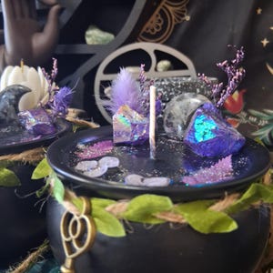 May include: A black cauldron candle featuring a dark wax surface topped with iridescent purple crystals, a grey moon-shaped stone, dried purple flowers, and glitter. The rim is wrapped in twine with green fabric leaves and a gold-toned skeleton key charm.
