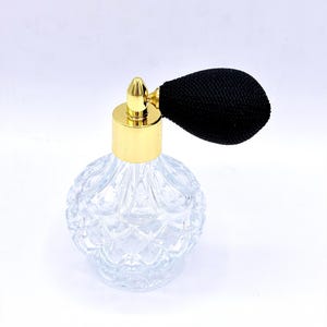 Puede incluir: Frasco de perfume de vidrio transparente con tapa dorada y bulbo atomizador negro. La botella tiene un patrón texturizado en forma de diamante. El atomizador está unido a la parte superior con una pieza metálica dorada.