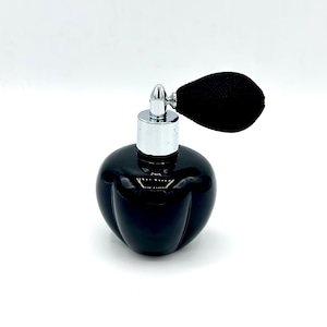 Puede incluir: Un frasco de perfume negro, redondo, con un atomizador plateado y un borla negra en forma de lágrima. El frasco tiene un acabado brillante y un diseño acanalado. El atomizador está centrado en el frasco.