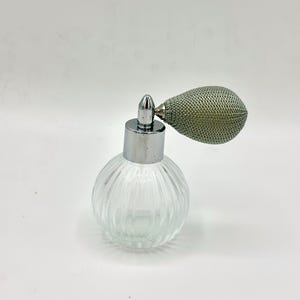May include: A clear, round glass perfume bottle with a ribbed design. It has a silver-colored metal collar and a bulb-shaped atomizer with a mesh covering. The bottle is empty and sits on a white surface.