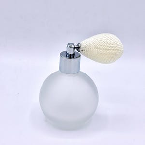 May include: A frosted glass perfume bottle with a silver metal top and a cream powder puff. The round bottle has a vintage design. The powder puff is attached to the bottle with a silver metal nozzle, a classic design.