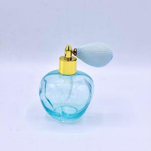 Puede incluir: Un frasco de perfume de estilo vintage con un cuerpo de vidrio azul claro y una tapa dorada. El frasco tiene una pera atomizadora azul claro a juego. El diseño recuerda a los frascos de perfume clásicos.