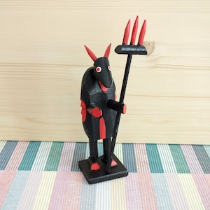 May include: A black wooden figurine of a devil with red horns and eyes, holding a black pitchfork with red tines. The figurine is standing on a black base.