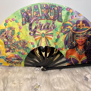 May include: A decorative hand fan with a black frame, featuring a vibrant Mardi Gras design. The fan displays the words "MARDI Gras" in sparkling letters, along with a woman in a mask and a fleur-de-lis. The color scheme includes green, purple, and gold.