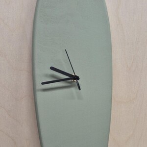 Op de afbeelding: Een surfboard-vormige wandklok in een gedempte groene kleur. De klok heeft zwarte wijzers en een minimalistisch ontwerp. De klok is gemonteerd op een lichtgekleurd houten oppervlak.