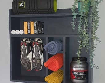 May include: A dark grey wall shelf with six compartments. The top compartment has a black and green foam roller. The second compartment has three white tubes. The third compartment has two black and white cycling shoes. The fourth compartment has a blue towel, a red towel, and a yellow towel. The fifth compartment has a black container with the text "Limited Edition SIS REGO Rapid Recovery" printed on it. The sixth compartment has a green plant hanging down.