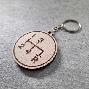 Puede incluir: Llavero de madera con un patrón de cambio de marchas grabado. El llavero presenta los números 1, 2, 3, 4 y R, junto con la palabra "Lift".