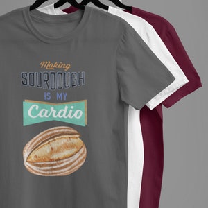 May include: Three t-shirts on hangers, the front one is gray with the text "Making sourdough is my cardio" and a drawing of a loaf of bread. The other two shirts are white and burgundy.