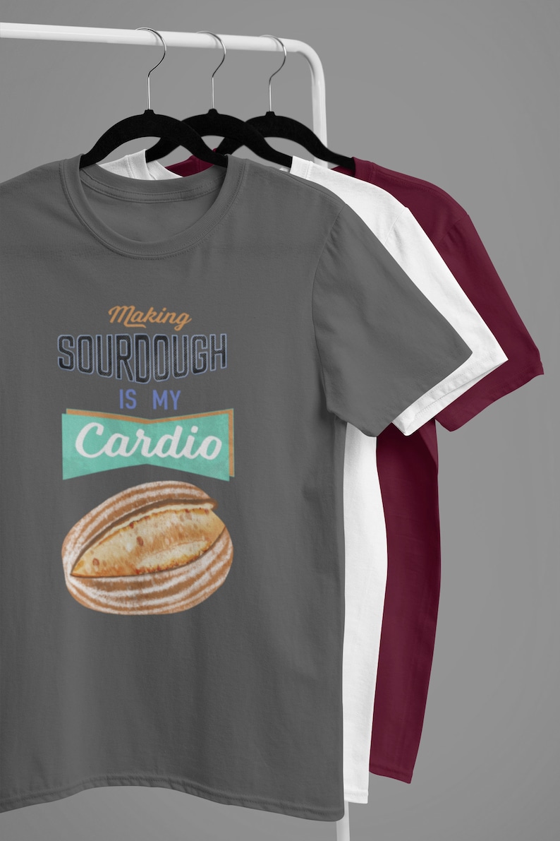 May include: Three t-shirts on hangers, the front one is gray with the text "Making sourdough is my cardio" and a drawing of a loaf of bread. The other two shirts are white and burgundy.
