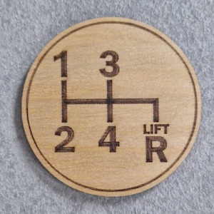 May include: A circular wooden coaster featuring a gear shift pattern. The numbers 1, 2, 3, 4, and R (reverse) are etched into the wood, along with the word "LIFT". The coaster is light brown with a dark brown etched design.