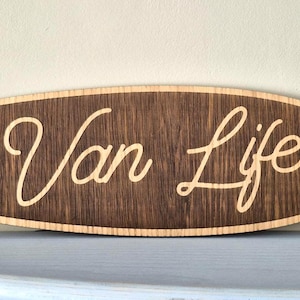 May include: A surfboard-shaped wooden wall sign with a dark brown wood grain finish. The text "Van Life" is engraved in a light-coloured script font in the centre, flanked by an engraved hibiscus flower on both the left and right sides.