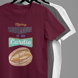 May include: Three t-shirts hanging on a rack. The front t-shirt is maroon and features a graphic of a loaf of sourdough bread with the text "Making Sourdough is my Cardio".