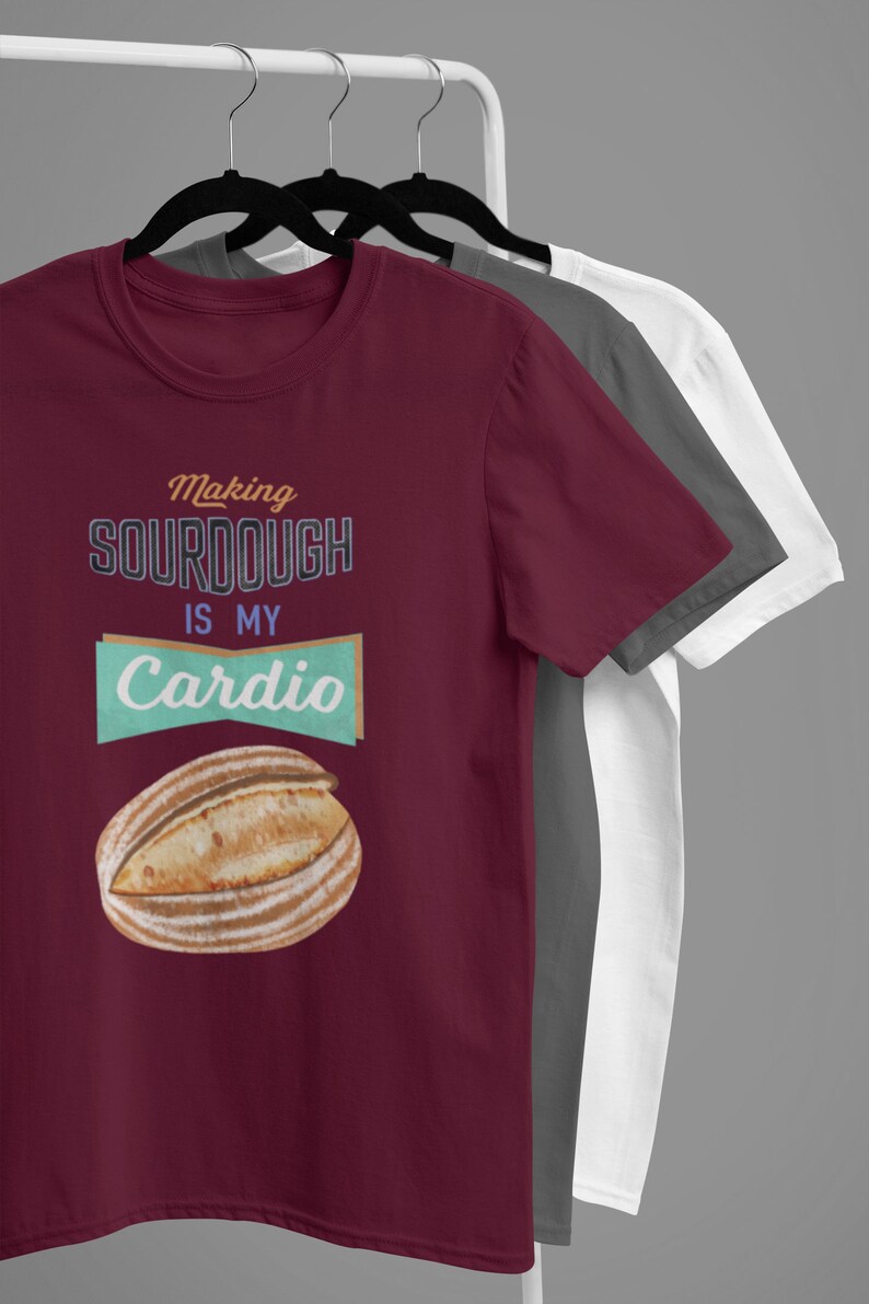 May include: Three t-shirts hanging on a rack. The front t-shirt is maroon and features a graphic of a loaf of sourdough bread with the text "Making Sourdough is my Cardio".