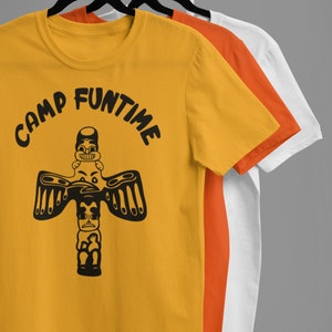 May include: Three t-shirts on hangers, the front one is yellow with a black graphic of a totem pole and the text "Camp Funtime". The middle shirt is orange and the back shirt is white.