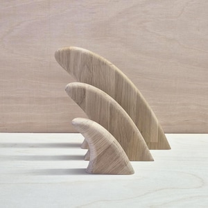 Puede incluir: Tres aletas de tabla de surf de madera de color claro dispuestas en orden descendente de tamaño, de mayor a menor. Cada aleta presenta una silueta curva y suave con un patrón de veta de madera natural visible, apoyada sobre una superficie.