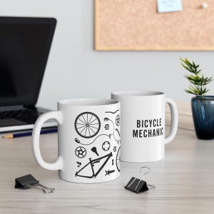 Può includere: Due tazze in ceramica bianca. Una tazza presenta illustrazioni nere di parti di biciclette. L'altra tazza ha il testo "BICYCLE MECHANIC" in nero. Le tazze sono su una superficie chiara.