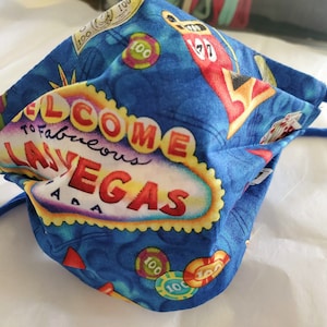 Może przedstawiać: Niebieska maseczka z tkaniny z motywem Las Vegas. Maseczka zawiera biały napis "Welcome to Fabulous Las Vegas" z czerwonymi, ż&oacute;łtymi i fioletowymi akcentami. Maseczka zawiera r&oacute;wnież obrazy kart do gry, kości i żeton&oacute;w do pokera.