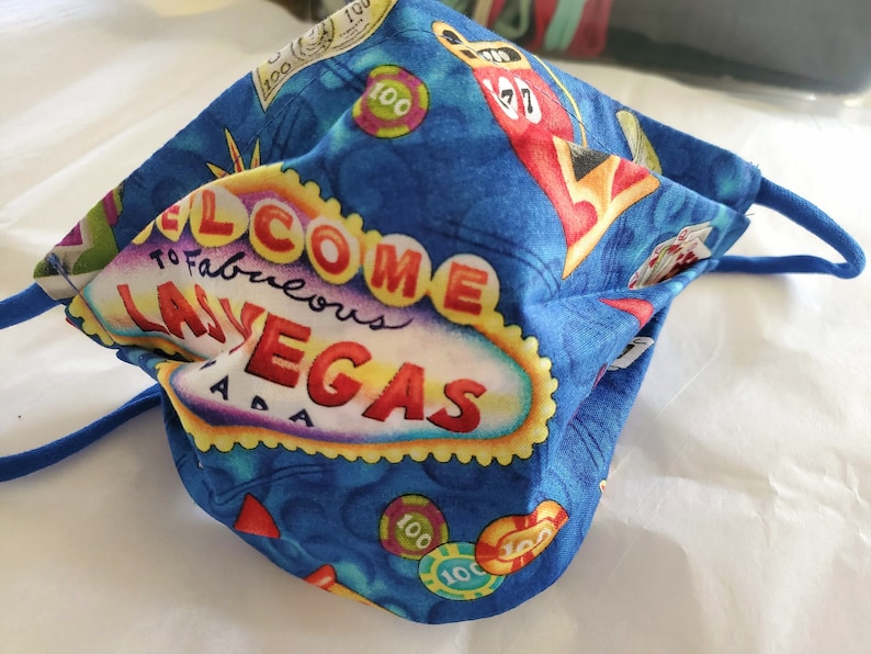 Może przedstawiać: Niebieska maseczka z tkaniny z motywem Las Vegas. Maseczka zawiera biały napis "Welcome to Fabulous Las Vegas" z czerwonymi, ż&oacute;łtymi i fioletowymi akcentami. Maseczka zawiera r&oacute;wnież obrazy kart do gry, kości i żeton&oacute;w do pokera.