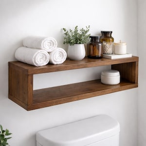 May include: A rectangular dark brown wooden floating shelf mounted on a white wall. The shelf holds three rolled white towels, a small potted green plant, two amber glass jars, a white candle, and a small white lidded container.