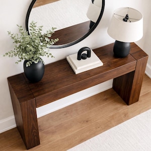 May include: A dark brown wooden console table with a black oval mirror, a black lamp with a beige lampshade, a black vase with greenery, and a decorative black and white object. The table is set against a white wall and a light brown wooden floor.