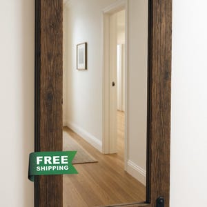 Peut inclure: Miroir rectangulaire avec un cadre en bois brun foncé et rustique. Le cadre présente un grain de bois visible et est orné de coins métalliques noirs. Le miroir est fixé sur un mur blanc, reflétant un couloir.