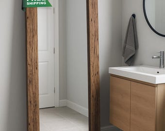 May include: A full-length rectangular mirror with a rustic, brown wooden frame. The mirror is leaning against a light grey wall in a bathroom setting. The text "FREE SHIPPING" is on a green banner attached to the frame. The sample colour is "Caskwood".