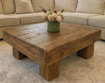 May include: A rustic, square coffee table made of reclaimed wood. The table has a natural wood finish and sits on four sturdy, block-style legs. A small, green metal container with dried flowers sits on top of the table.