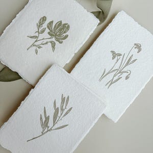 May include: Three white handmade paper cards with botanical illustrations in olive green. Each card features a different floral or plant design, including a flower, sprigs, and leaves. The cards have deckled edges.